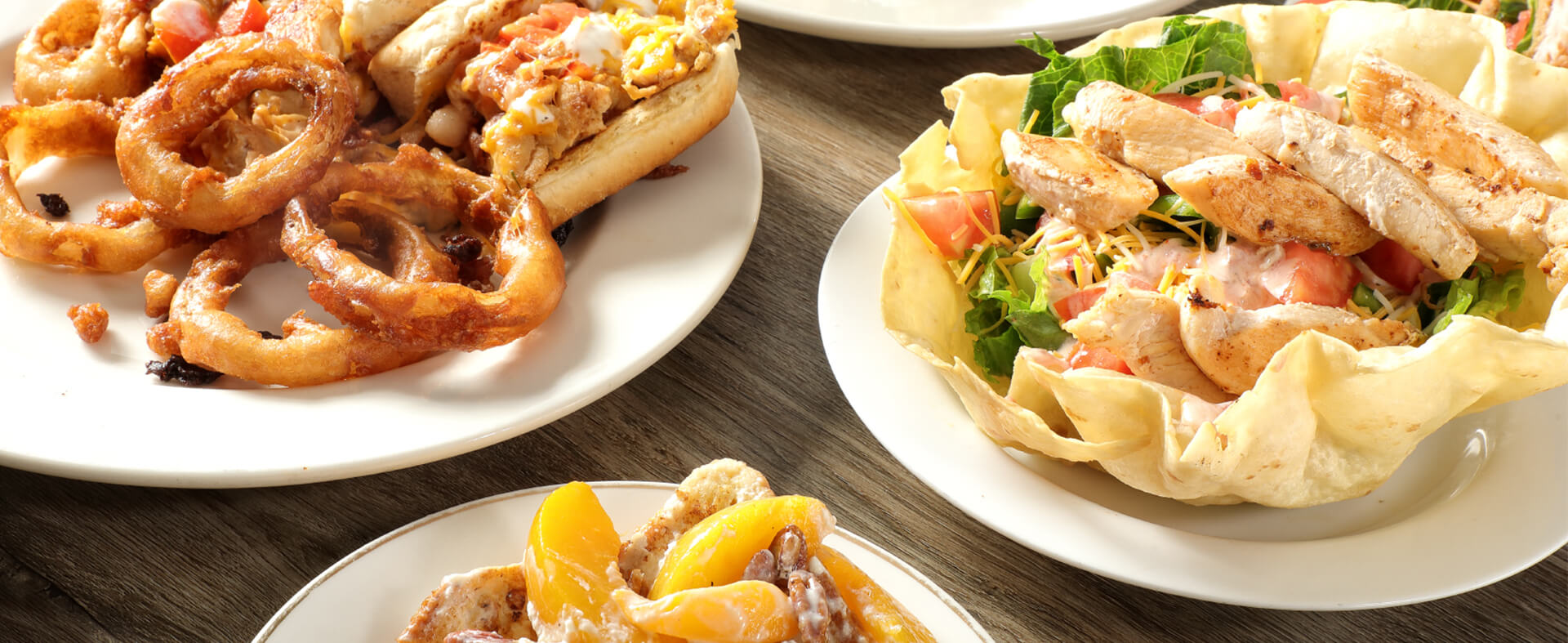 A close-up of chicken sandwiches, onion rings, and chicken salad in tortillas is on white plates. This shows the menu variety