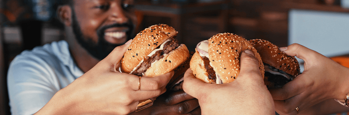 Three different ethnic groups are holding up their burgers in the air. They are enjoying lunch together.