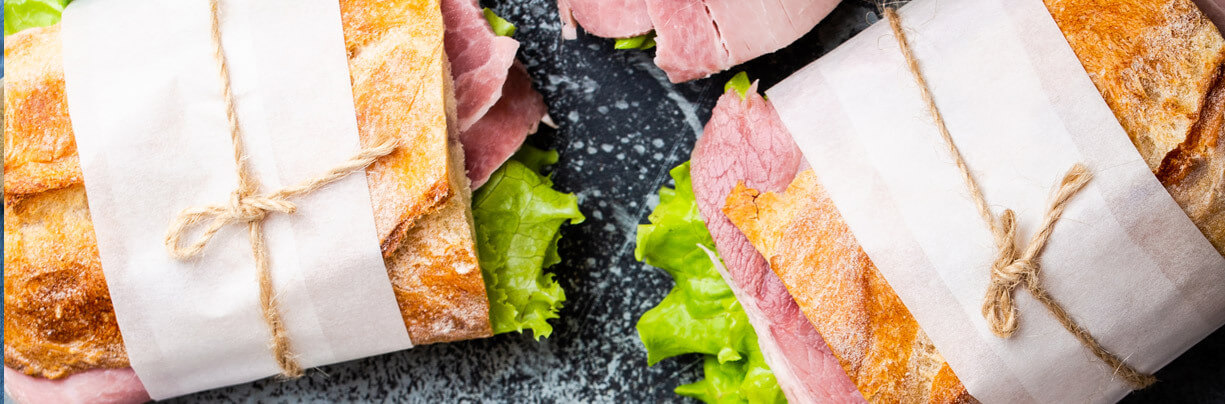 Three wrapped ham sandwiches with lettuce on ciabatta bread, tied with string on a gray stone background