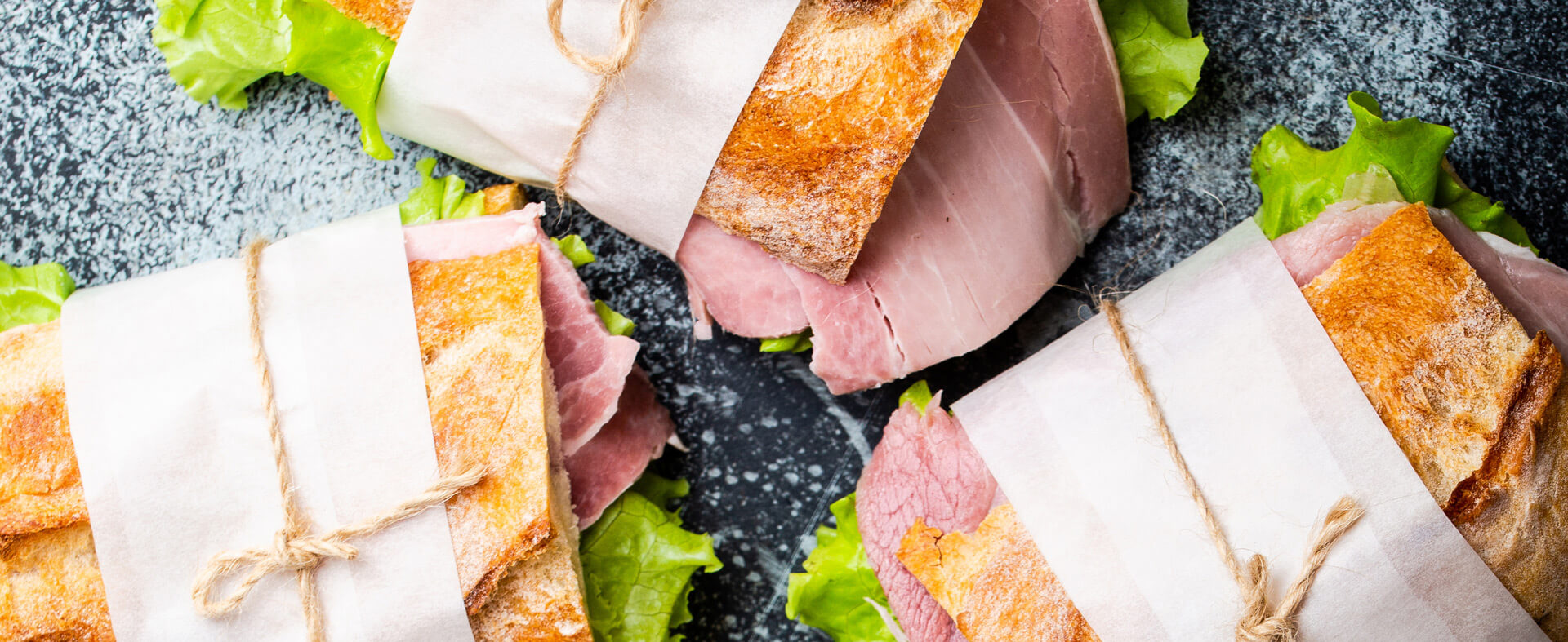 Group of three wrapped deli sandwiches tied with string on a gray stone background.