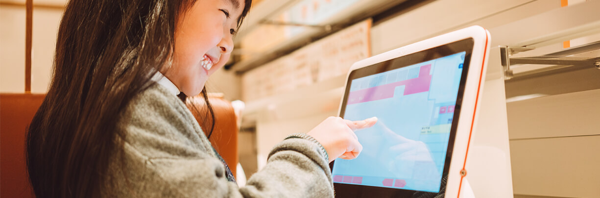 A cute, smiling young Asian girl pointing at a food item she wants on a touch screen kiosk.