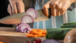 Close-up of male hands thinly slicing red onion using a chef knife on a cutting board.