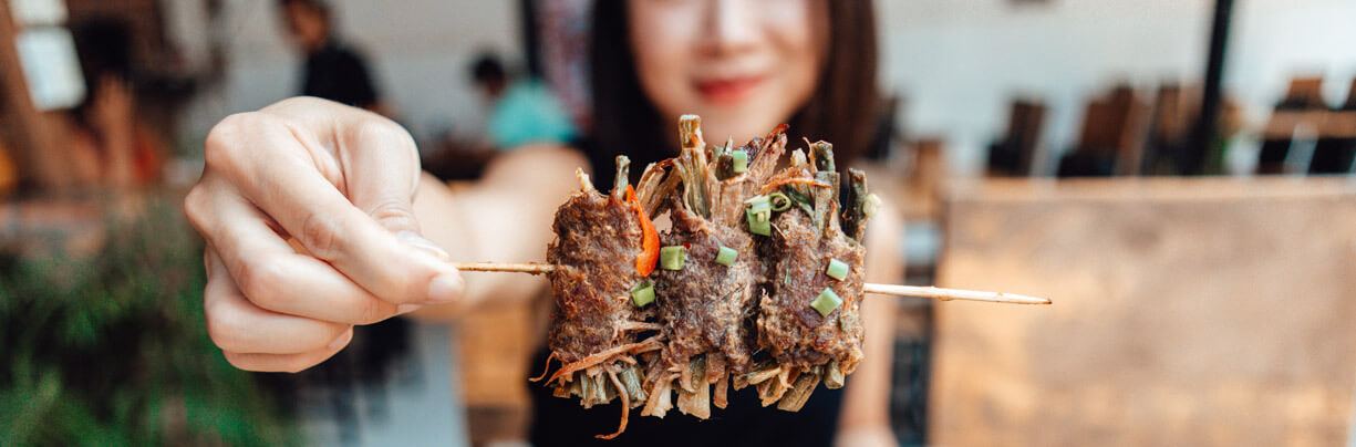 Close-up image of an authentic Thai beef skewer in the hand of an attractive young Asian female.
