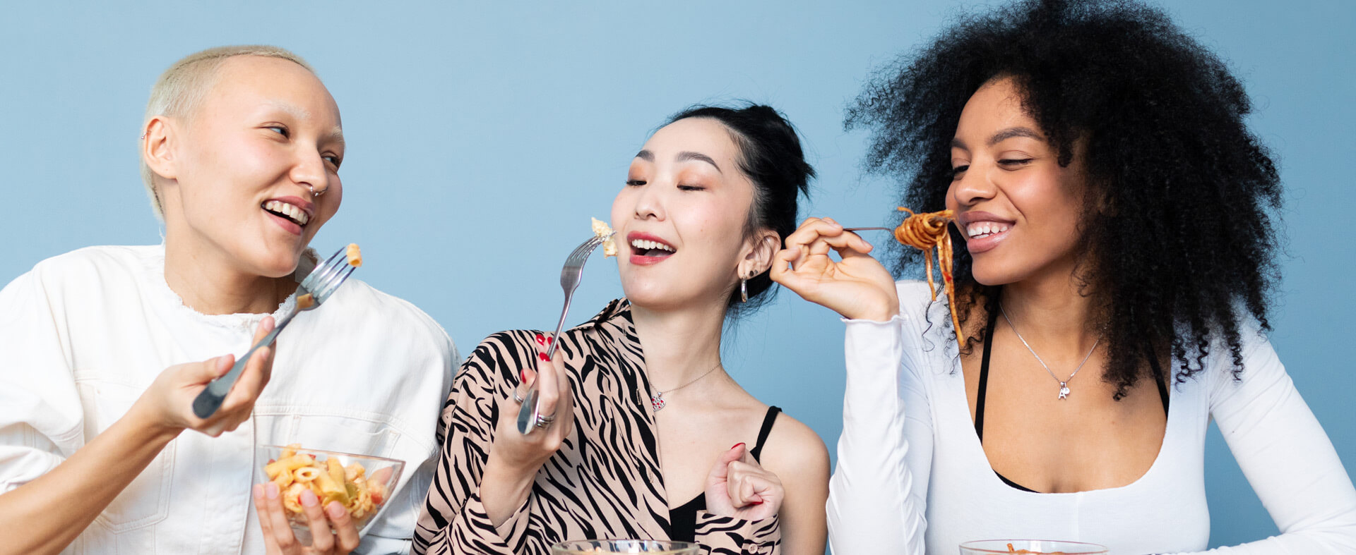 Three ethnically diverse female Gen Z friends eating pasta with a fork.