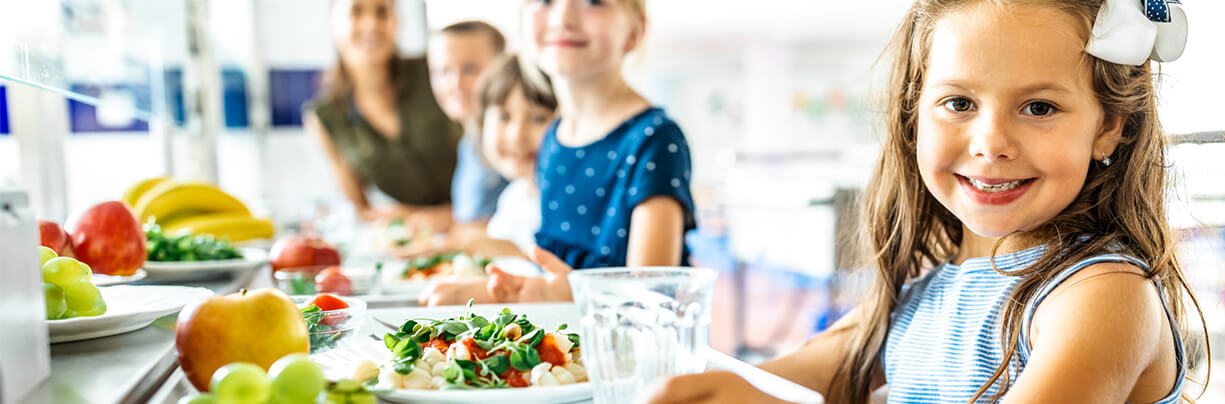 Five K-12 kids in a lunch line putting healthy food on trays with one girl in focus and the others out of focus.