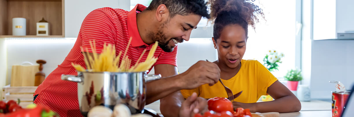 Happy black father and daughter cutting tomatoes and preparing a healthy pasta meal in their home kitchen.