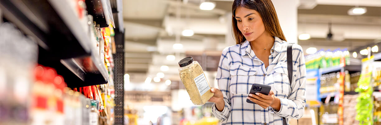 Young female shopper in a convenience store searching on her phone for product information while holding a spice container.