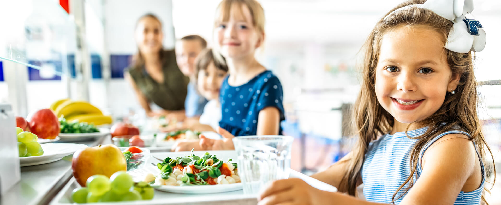 Close up of a young girls holding a school lunch tray of healthy food with classmates in a row at the lunch line.