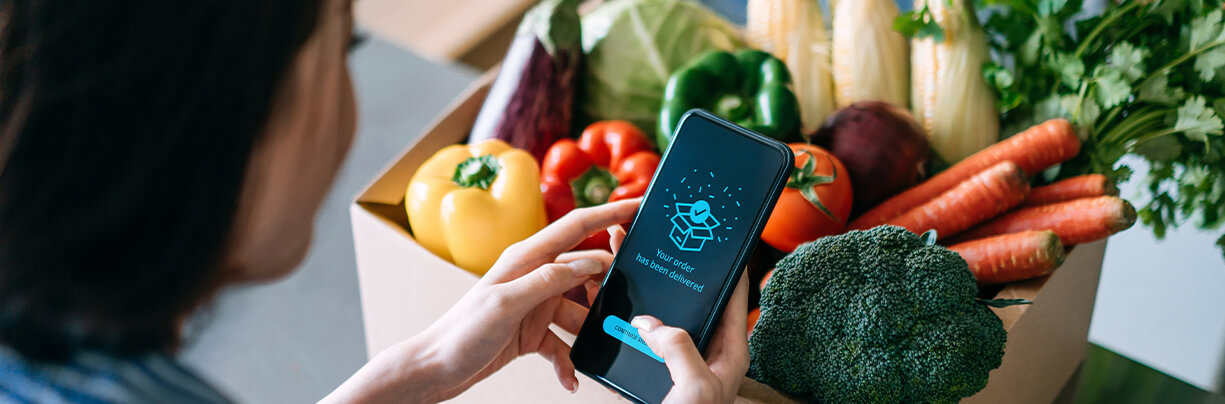A woman is holding her phone, looking at her online delivery phone app after her box of fresh vegetables.