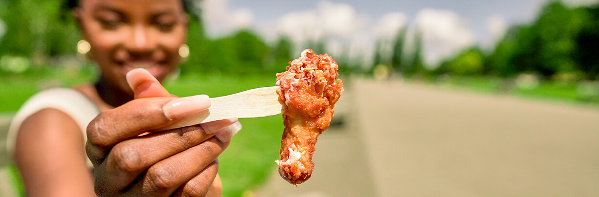 Close-up of an African American Gen Z female's hand holding a crispy chicken wing with a wooden to-go fork.
