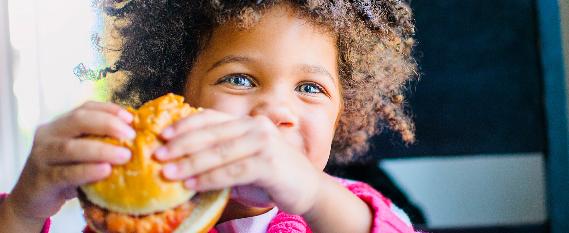 A closeup of an adorable African-American little boy smiling and holding a chicken sandwich at a restaurant.