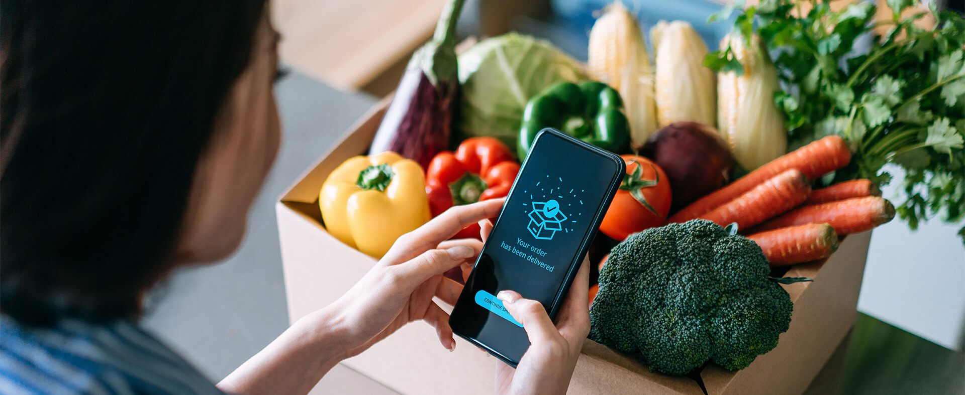 Asian woman holding her phone with an app indicating her grocery order has been delivered with a box of vegetables.