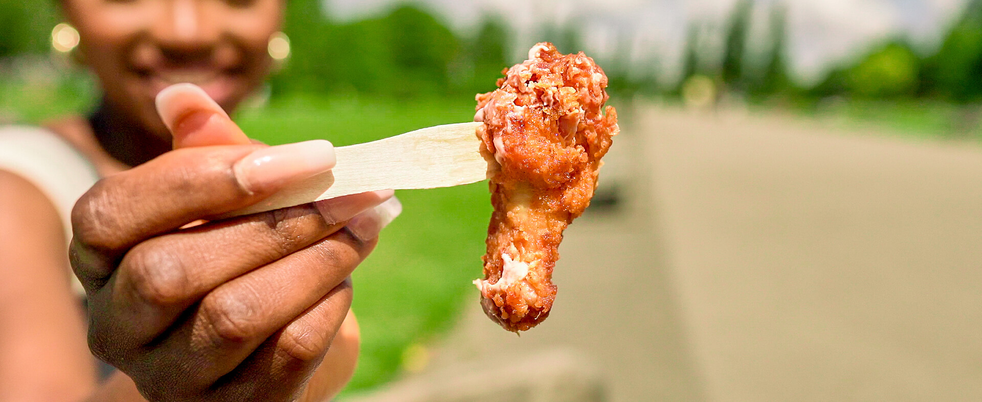 Young black female holding onto a breaded chicken wing with a fork, eating her take-out fried chicken wing order on a bench out in the sunshine.