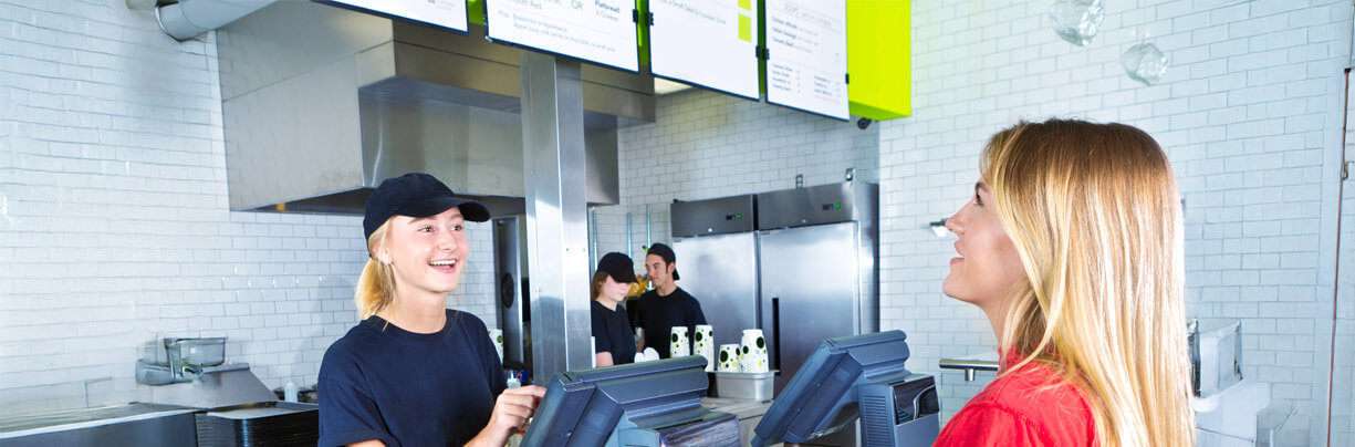 A teenage female standing behind a counter at a fast food restaurant takes an order from another female who is reading an overhead menu board.