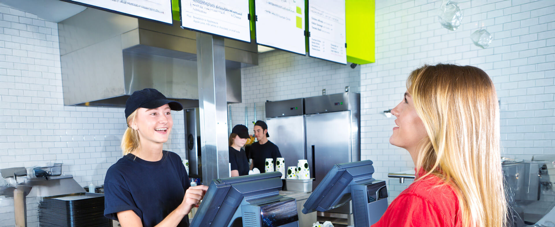 Young female working at a fast food restaurant takes an order from a 20-something female looking up at a menu board.