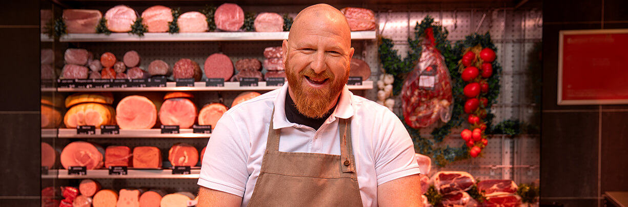 Image of the inside of a butcher shop with a happy male butcher at his counter and a large meat display behind him.