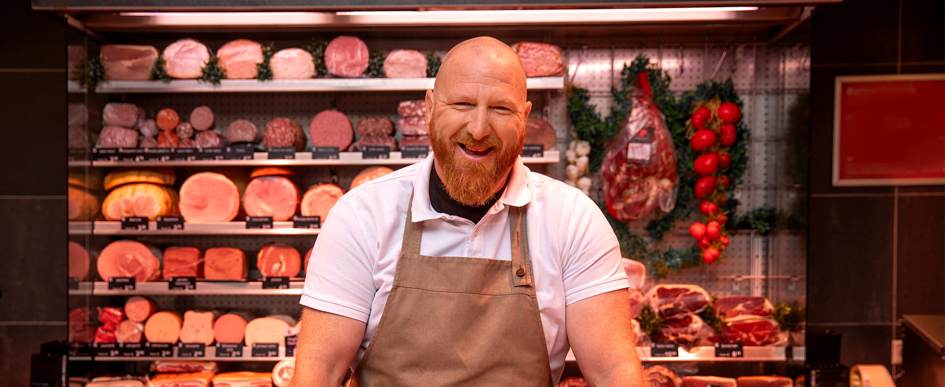 Smiling male butcher at his counter in front of various types of meat on display.
