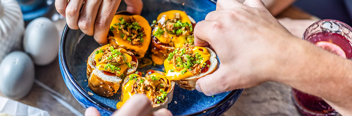 Group of hands each pulling a sample of an appetizer of a baguette with meat and cheese.