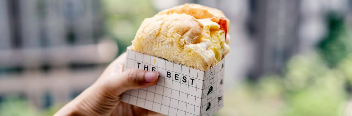 A handheld breakfast sandwich in a cardboard container is being held by a single hand while outdoors.