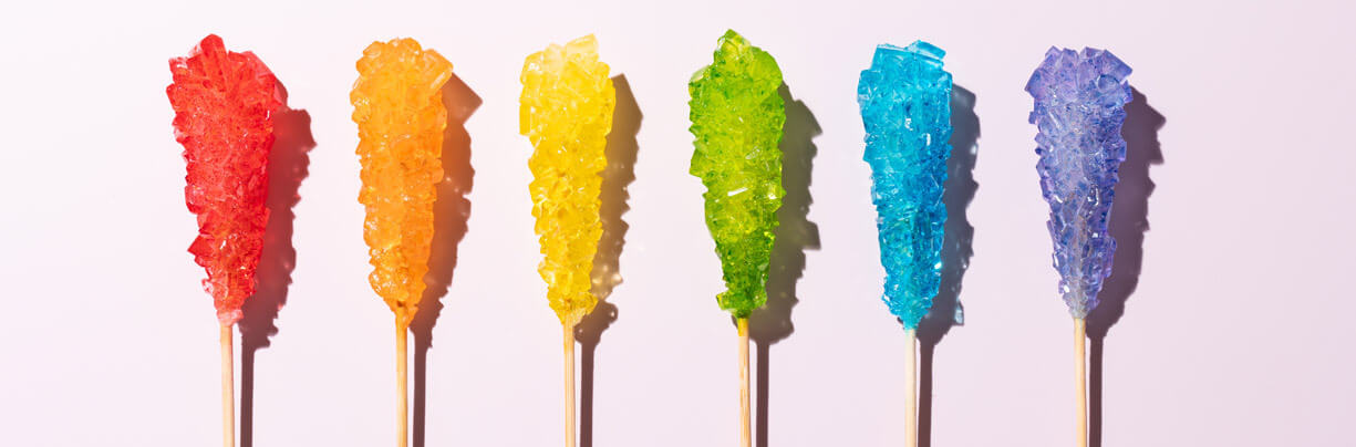 Six different colors of rock candy on a stick in a row on a pink background.