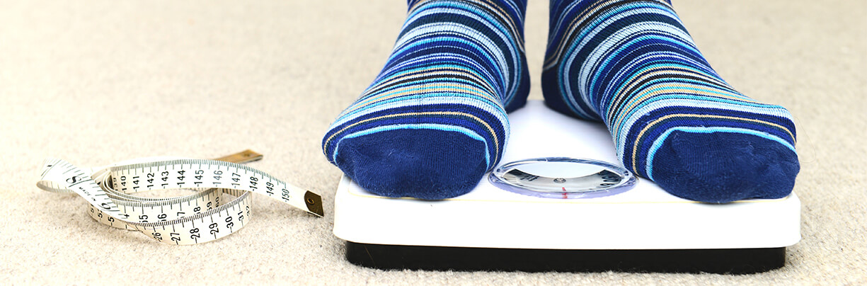 A front-on close-up image of human feet wearing socks and standing on a scale next to a curled-up measuring tape.