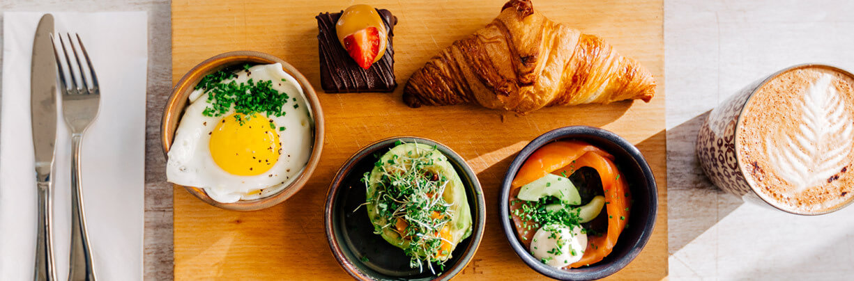 Cutting board with small serving cups filled with foods like a fried egg, avocado, peppers, croissant and a strawberry-topped chocolate piece.