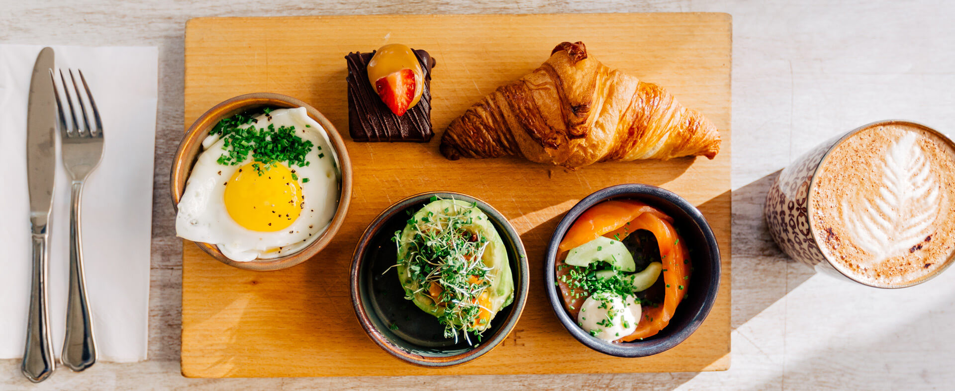 A horizontal image of a group of foods in small portion containers lying on a cutting board with silverware on the left and a cup of cappuccino on the right.