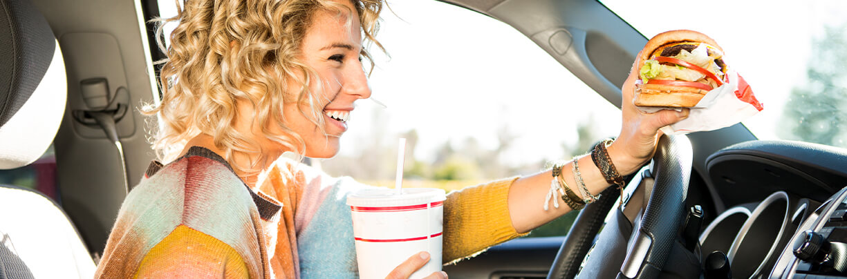 A Caucasian young adult driving in her car, eating a burger and drinking a beverage from a fast food place.