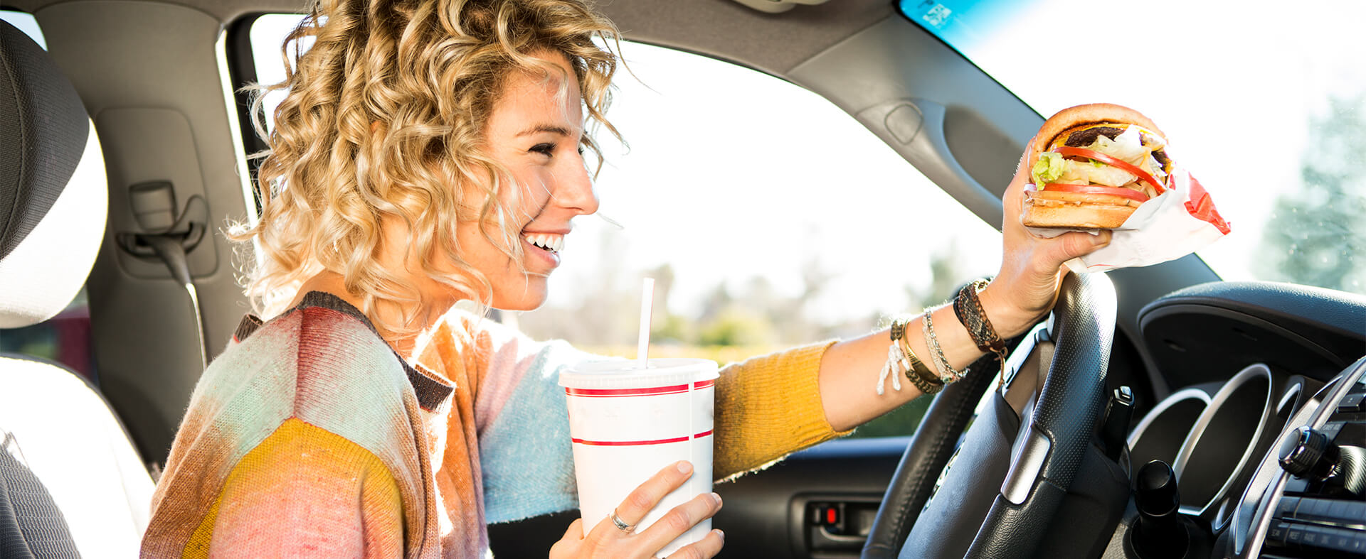 Young female eating a fast-food burger and beverage in her car while driving.