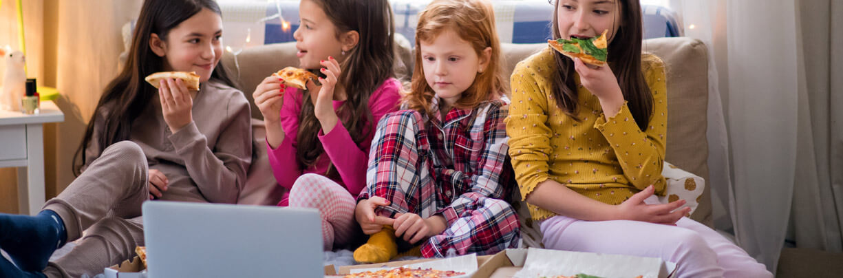 Four Gen Alpha girls at a slumber party, sitting on a bed, eating pizza and watching a laptop.
