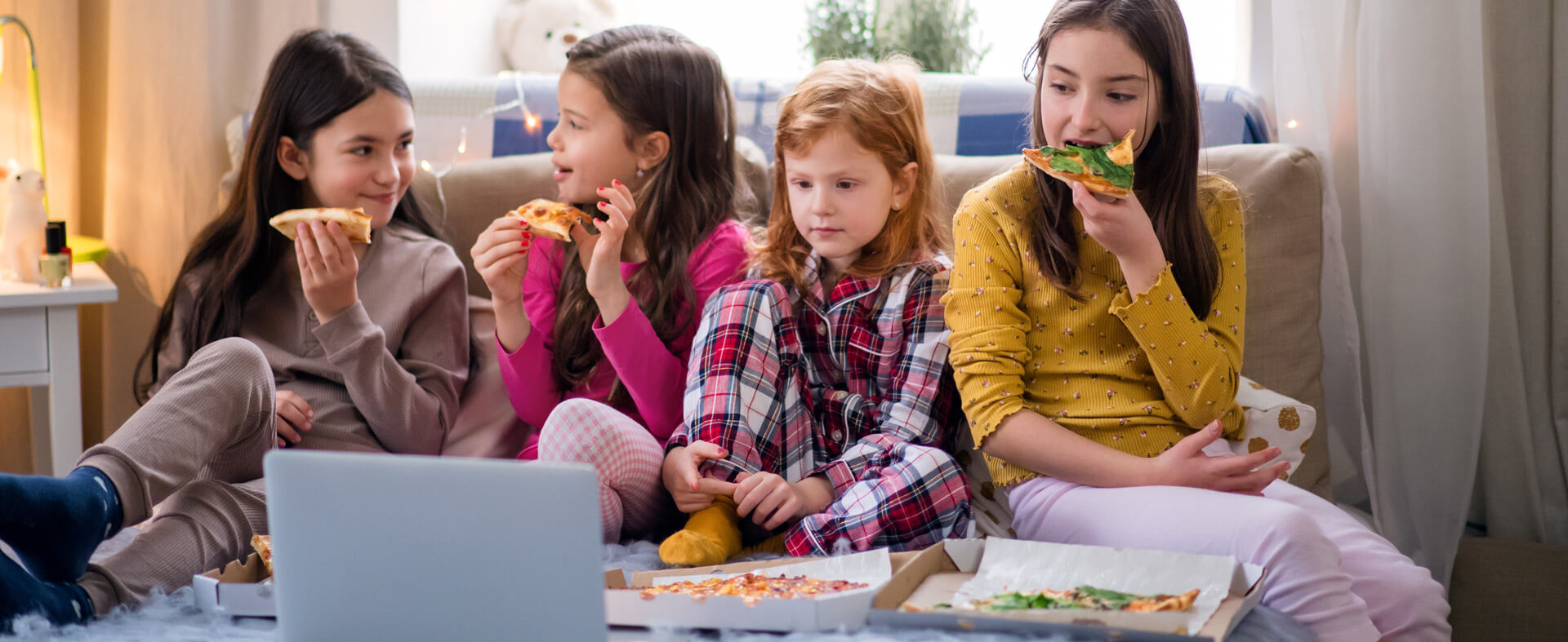 Four Gen Alpha girls sitting on a bed, eating pizza and watching a laptop.