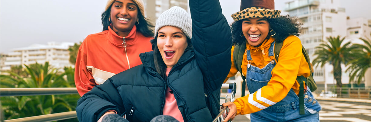 Two Gen Z women of different ethnicities pushing another in a shopping cart while happily running through a supermarket parking lot.