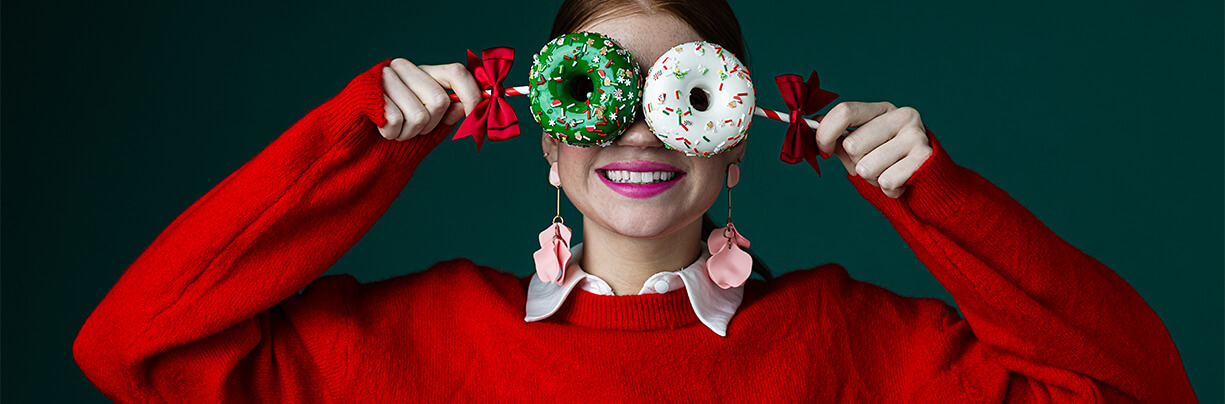 Young female in a red sweater holding up two holiday-themed donuts over her eyes.