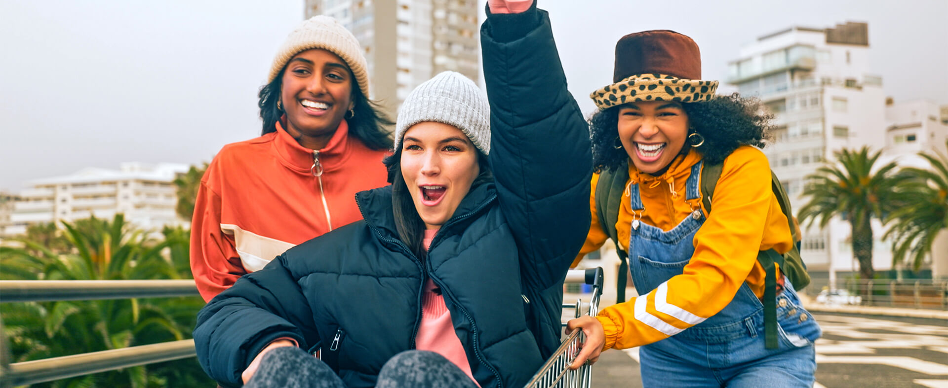 Three Gen Z women of different ethnicities running through a supermarket parking lot in a shopping cart.