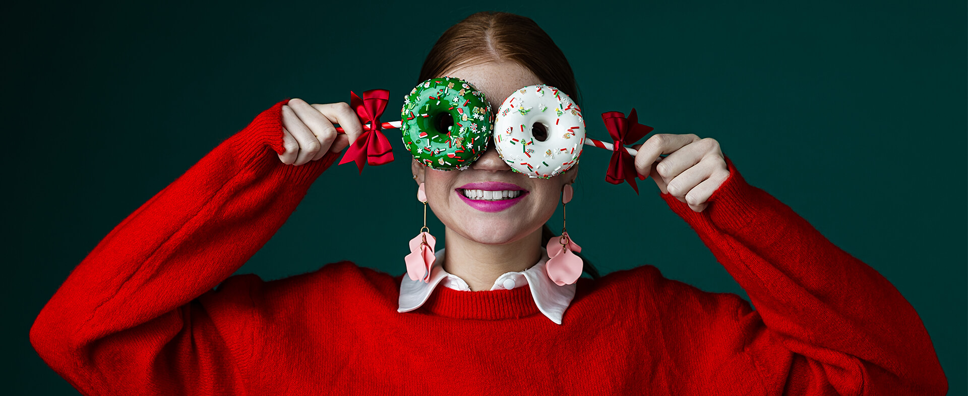 Gen Z female in a red holiday sweater, covering her eyes with two festive, decorated donuts.