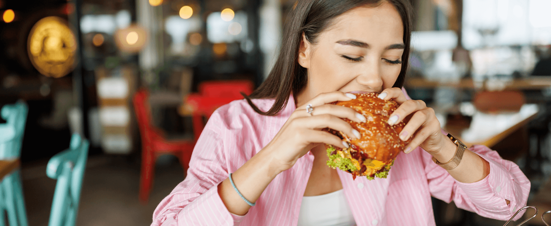 Close-up image of a Gen Z female taking a bite out of a burger in a casual dining restaurant with a blurred background.