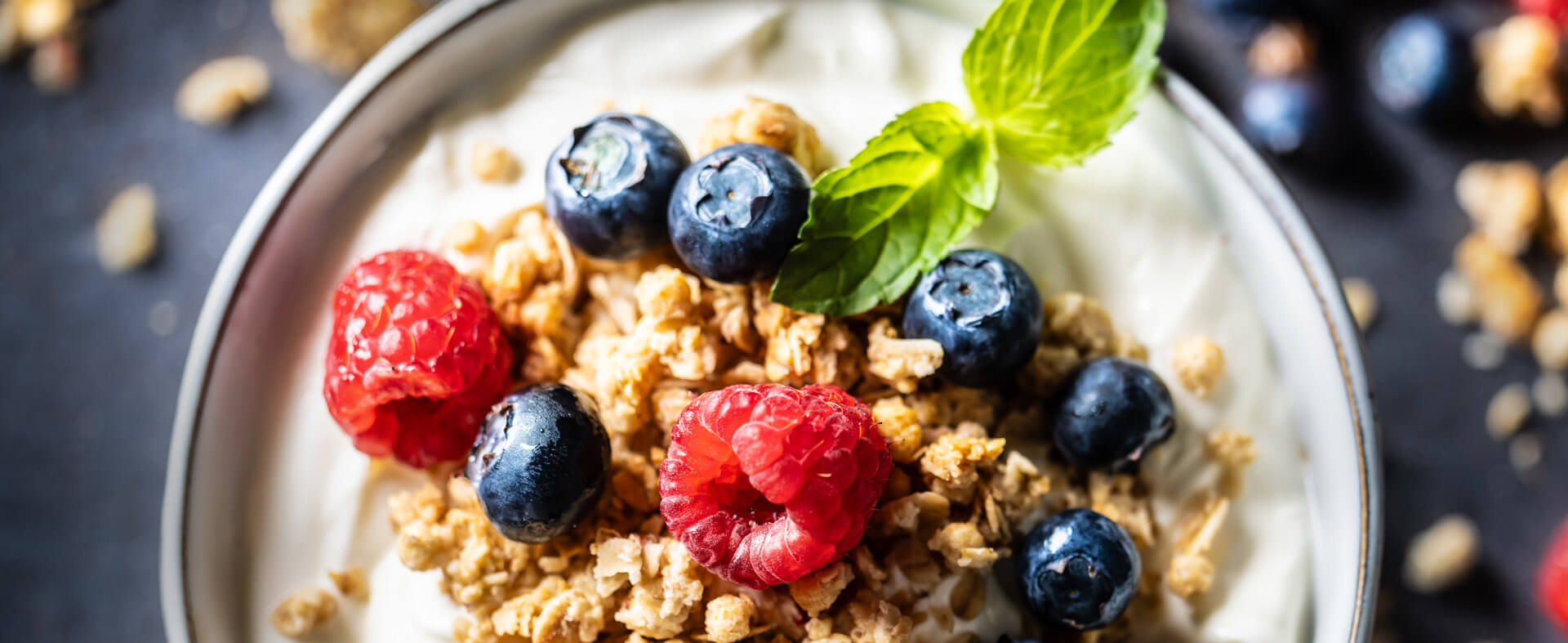 Top-down image of a bowl of Greek yogurt with oats, raspberries, and blueberries, with ingredients spilled around it and out of focus.