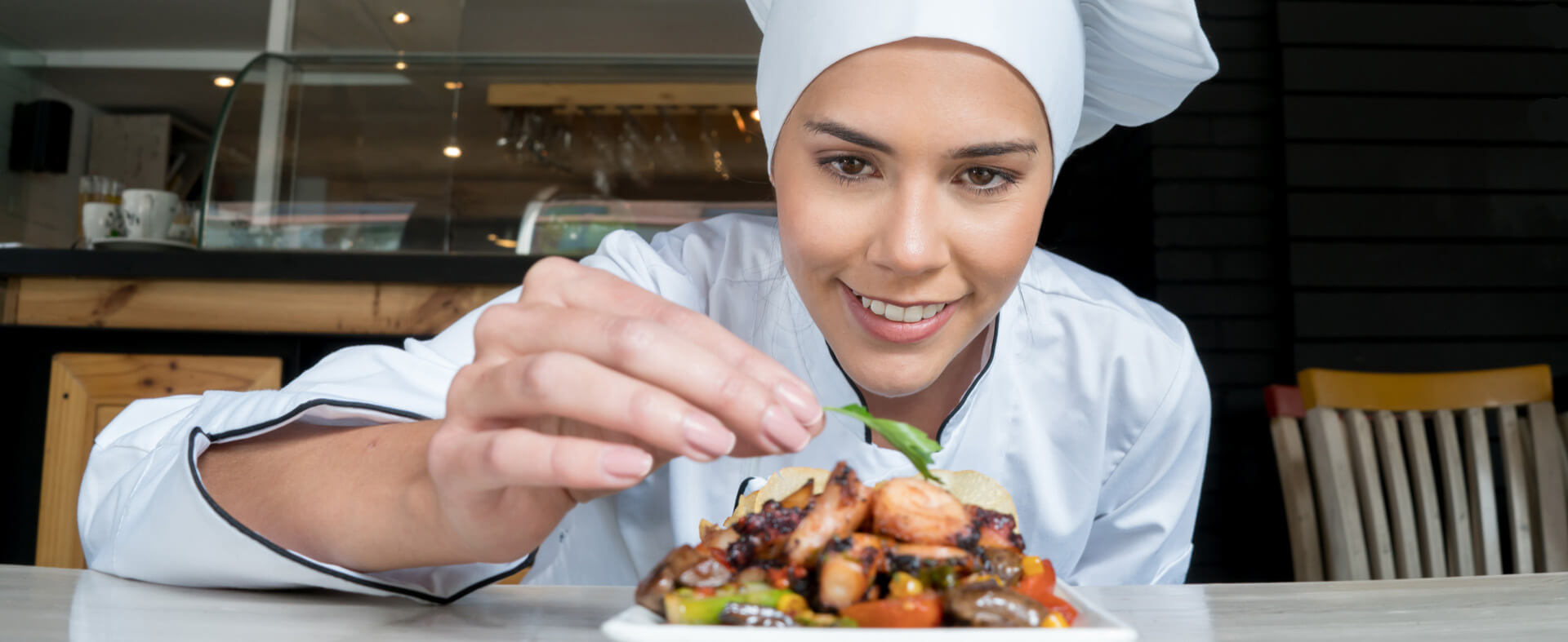 A happy female chef is putting the finishing touches on an entree by placing a piece of lettuce on top.