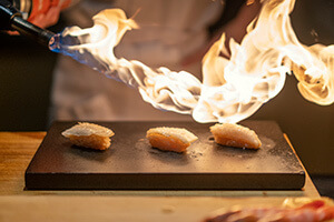 Image of a chef heating up three pieces of sushi with a torch and flame.