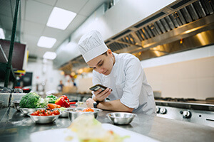 A male chef in the kitchen of a restaurant is looking for a recipe on his phone.