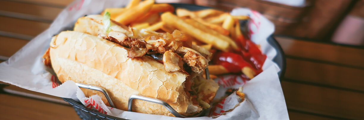 Close-up image of a spicy chicken sub sandwich with fries and ketchup the background, sitting in a plastic basket showing ballpark food.