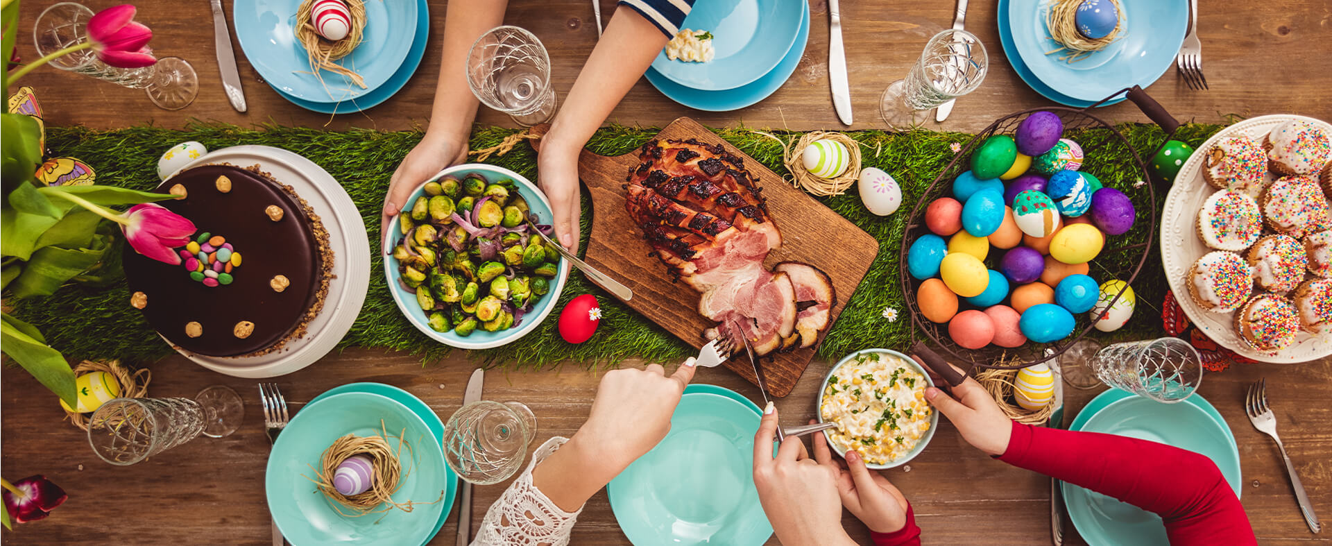 Overhead shot of an Easter table with ham, veggies, Easter eggs and desserts being served and passed around by hands.
