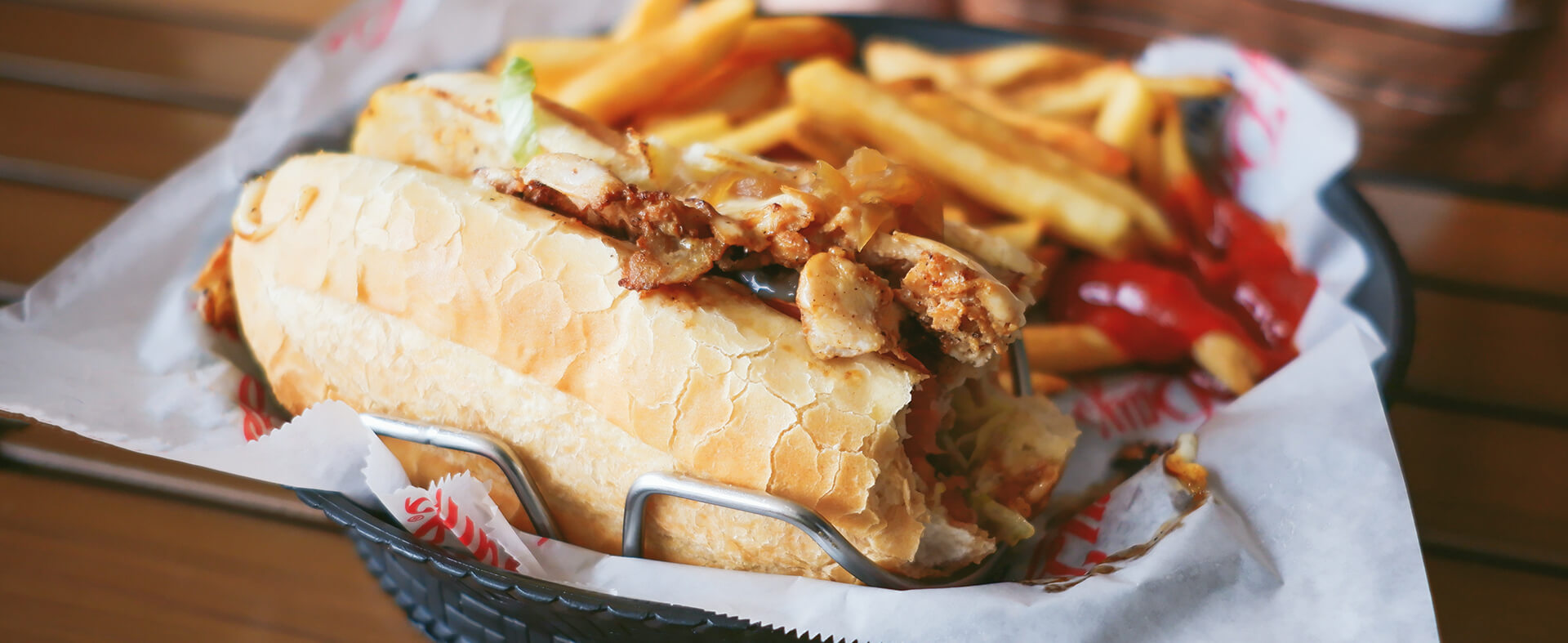 Enlarged image of a chicken sub sandwich with fries and ketchup in a plastic basket showing ballpark food.
