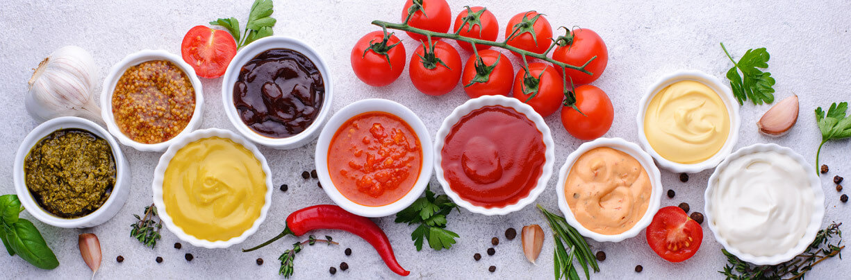 A straight down shot of a grouping of different flavored sauces in white ramekins with ingredients around them.