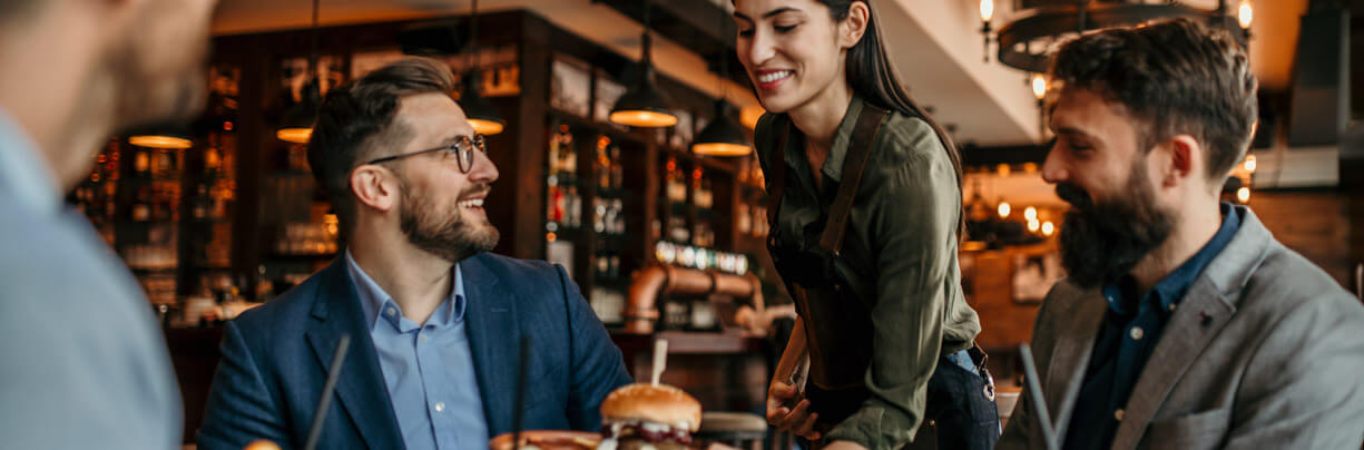 In a bar and grill restaurant, a friendly female server delivers a plated burger to a table of business casual men.