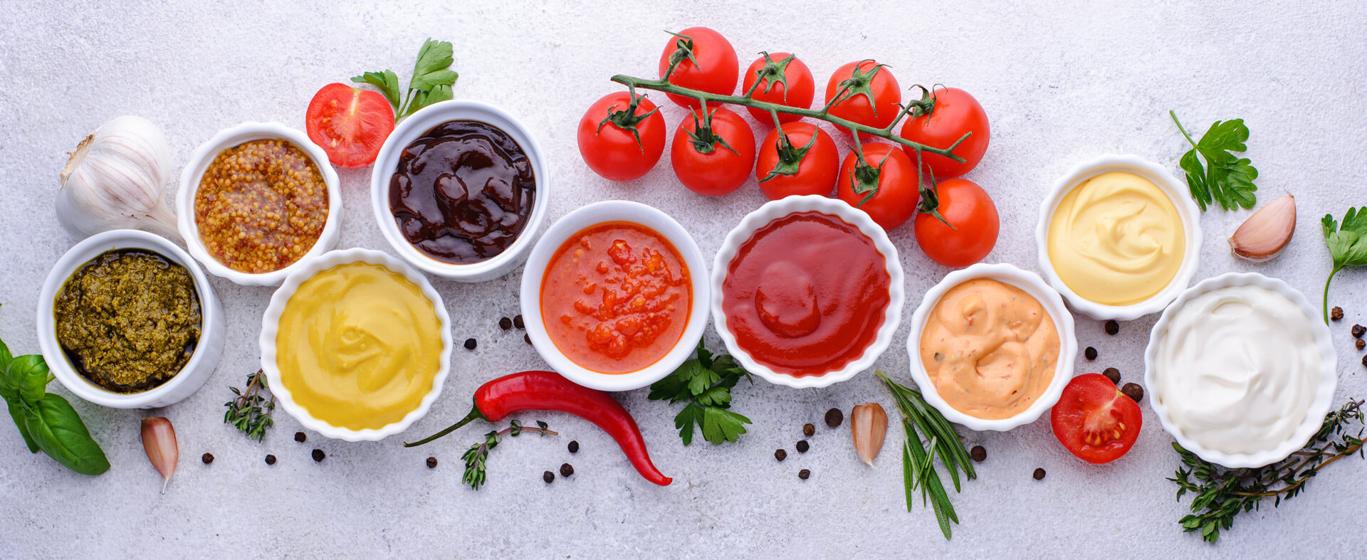An overhead shot of various sauces in white containers in a row, with herbs and vegetables scattered around.