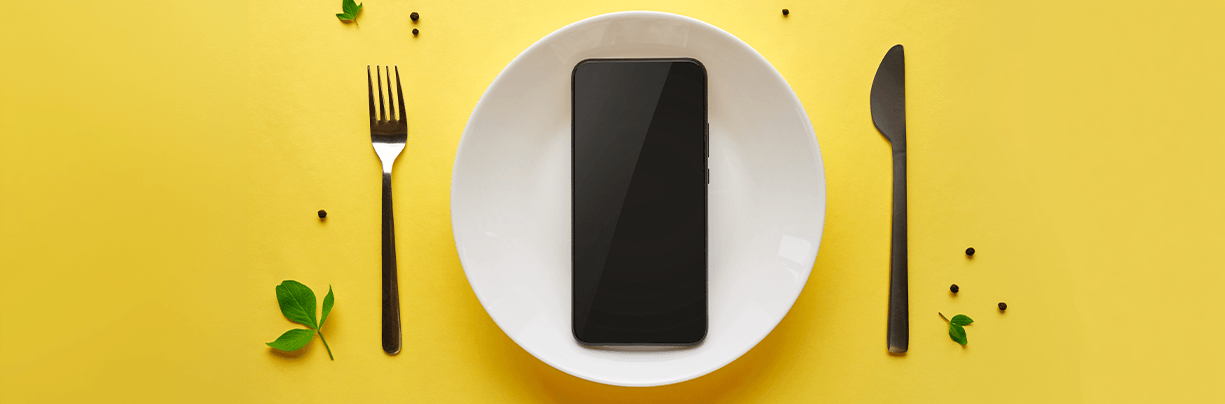 Smartphone on a white plate, with the fork and knife on each side, on a yellow background.